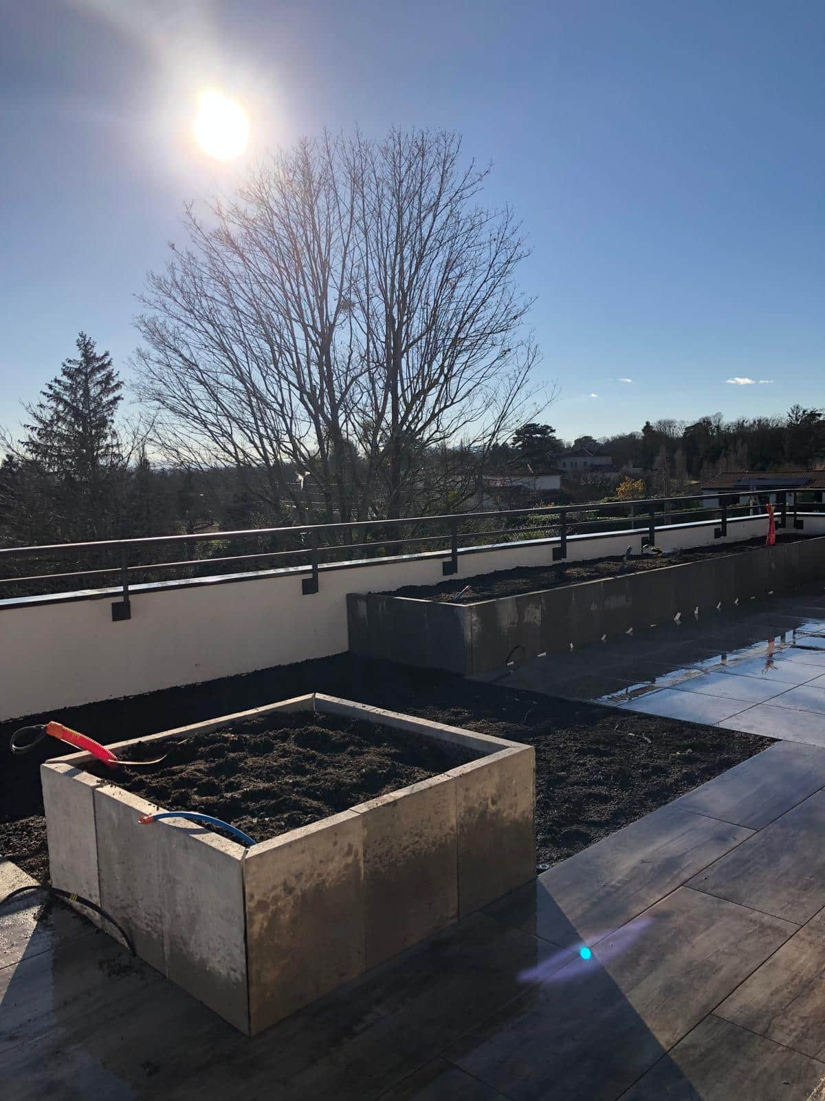 Terrasse ensoleillée avec jardinières en béton remplies de terre et dalles grises, vue sur arbres nus et ciel bleu.