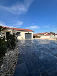Allée de maison moderne avec pavés foncés, garnie de gravier blanc et plantes vertes, sous un ciel bleu clair.