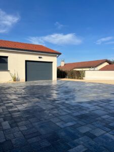 Allée en pavés gris foncé texturés devant un garage moderne, sous un ciel bleu et ensoleillé.