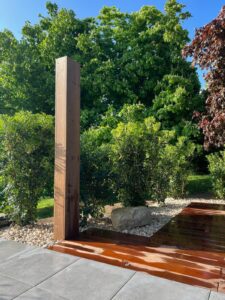 Structure en bois brun devant des arbustes verts, près d'une terrasse en dalles grises et planches luisantes.