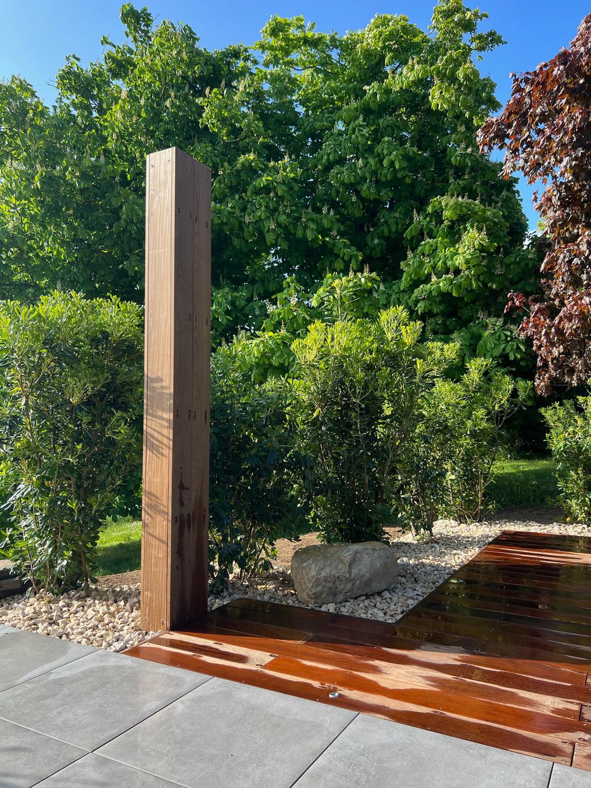 Structure en bois brun devant des arbustes verts, près d'une terrasse en dalles grises et planches luisantes.