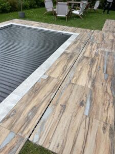 Margelle de piscine carrelée aspect bois clair et gris, avec couverture de sécurité et drain linéaire au sol.