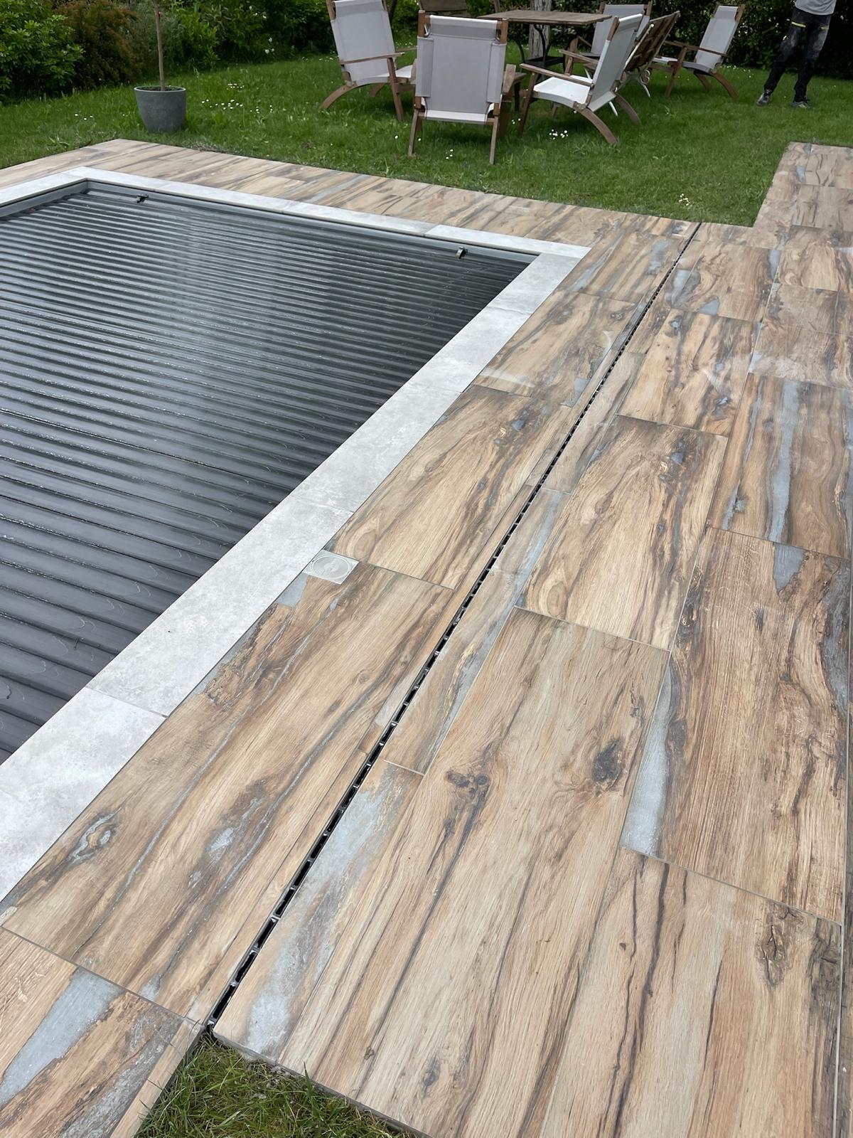 Margelle de piscine carrelée aspect bois clair et gris, avec couverture de sécurité et drain linéaire au sol.