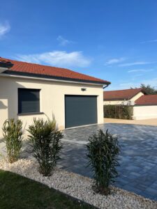 Façade d'une maison moderne avec toit en tuiles rouges, porte de garage gris anthracite, volet roulant fermé et aménagement paysager de gravier blanc et plantes vertes.