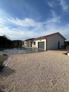 Maison individuelle moderne avec toit tuiles rouges, garage gris et allée gravier clair devant une zone pavée sombre sous ciel bleu.