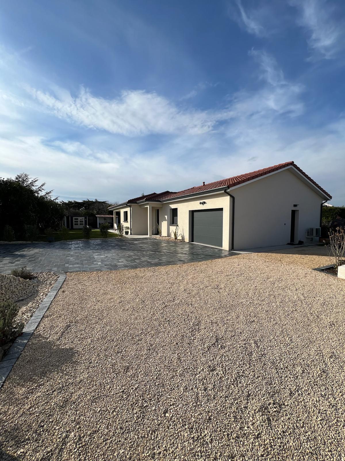 Maison individuelle moderne avec toit tuiles rouges, garage gris et allée gravier clair devant une zone pavée sombre sous ciel bleu.