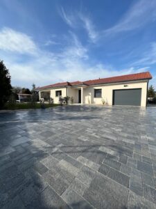 Maison moderne de plain-pied avec toiture tuiles rouges et grande allée pavée gris foncé sous un ciel bleu nuageux.