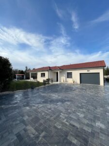 Maison plain-pied moderne avec toit tuiles rouges, façade crème, et grande allée en pavés gris foncé sous un ciel bleu et nuageux.