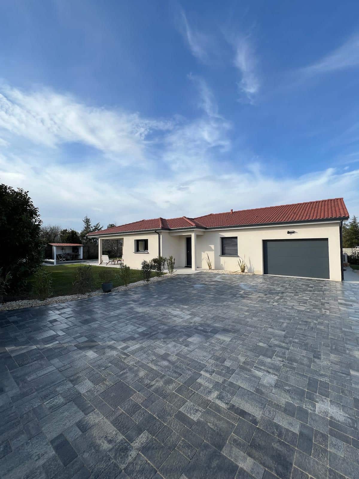 Maison plain-pied moderne avec toit tuiles rouges, façade crème, et grande allée en pavés gris foncé sous un ciel bleu et nuageux.
