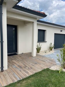 Façade d'une maison moderne avec terrasse en carrelage imitation bois, porte d'entrée anthracite et garage, sous un ciel nuageux.