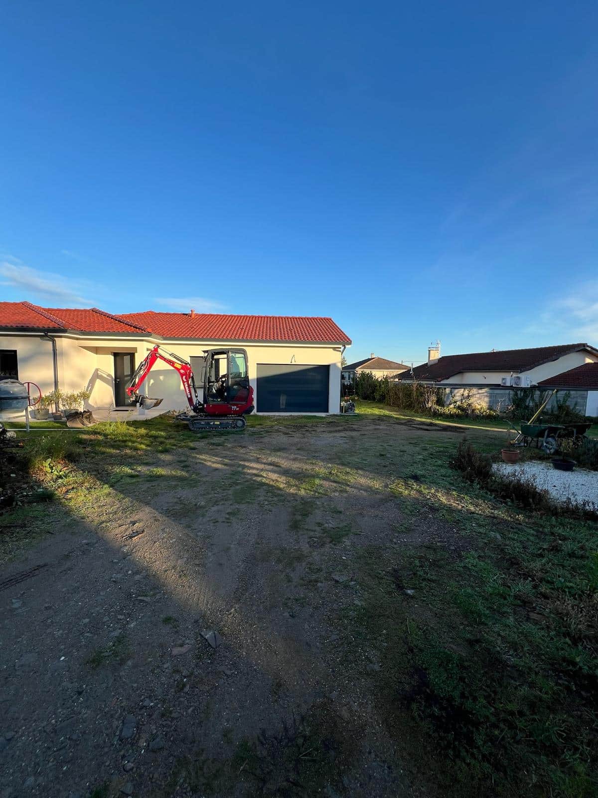 Mini-pelle rouge Yanmar 7T en chantier devant une maison claire avec toit tuiles rouges et ciel bleu clair.