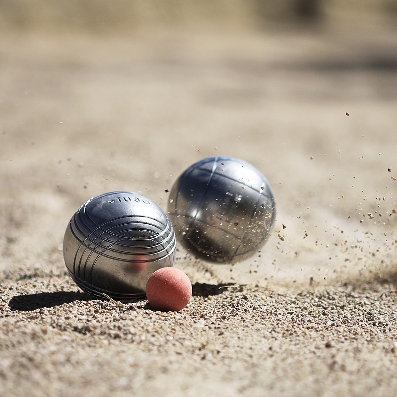 Boules de pétanque en action. Deux boules argentées et un cochonnet rouge sur gravier, l'une "TUBO" frappant le sol avec des projections.