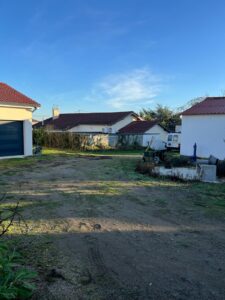 Terrain dégagé avec pelouse et terre battue, maisons à toits rouges, ciel bleu clair et soleil bas.