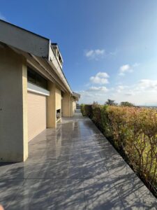 Allée carrelée grise et brillante longeant une façade beige avec volets roulants, sous un ciel bleu parsemé de nuages.