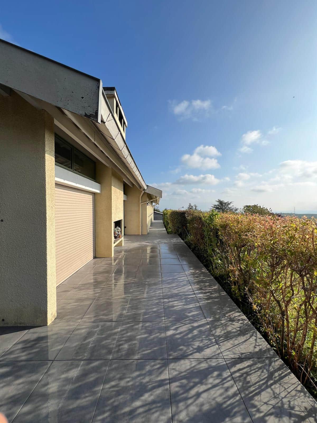 Allée carrelée grise et brillante longeant une façade beige avec volets roulants, sous un ciel bleu parsemé de nuages.