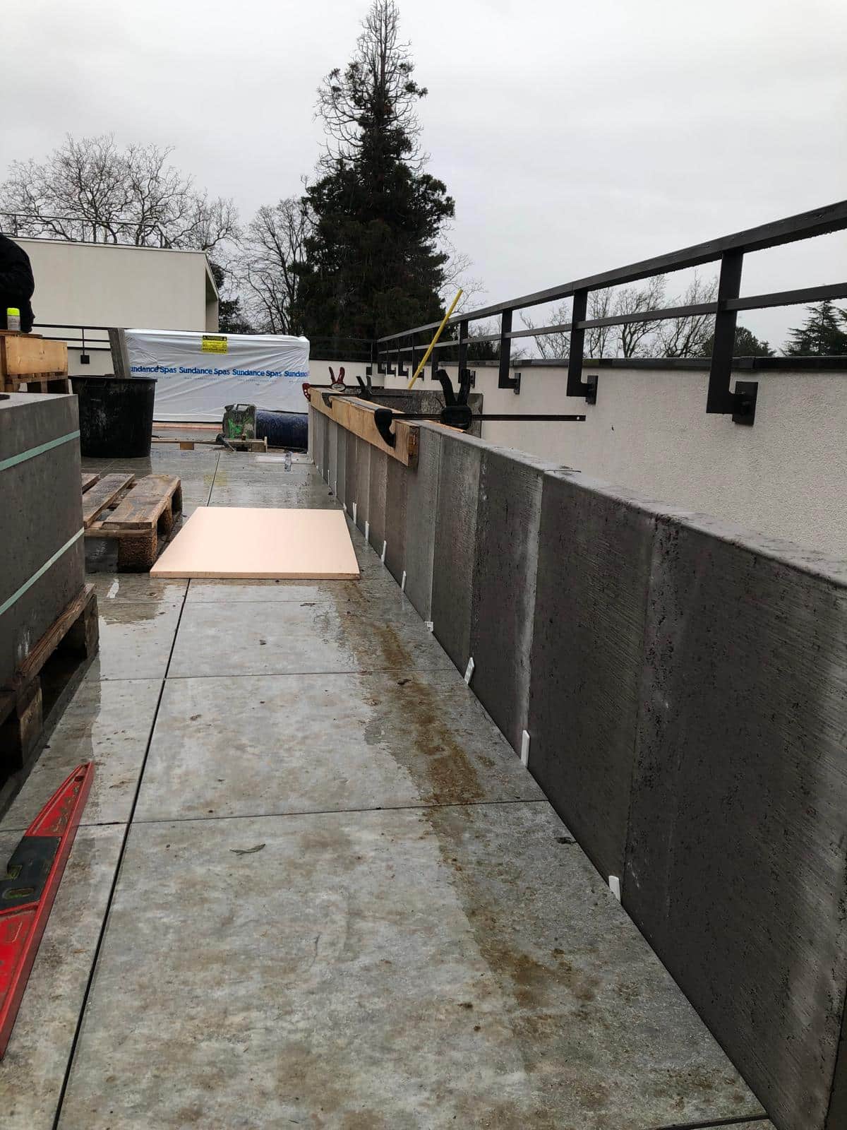 Chantiers terrasse en travaux : blocs de béton, palettes, mousse isolante rose, rambarde noire, ciel gris et grand arbre au loin. Sundance Spas visible.