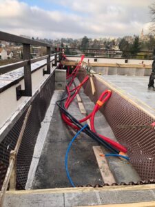 Chantier de terrasse en cours : gaines rouges et noires, drainage alvéolaire, et garde-corps en métal avec vue sur ville.