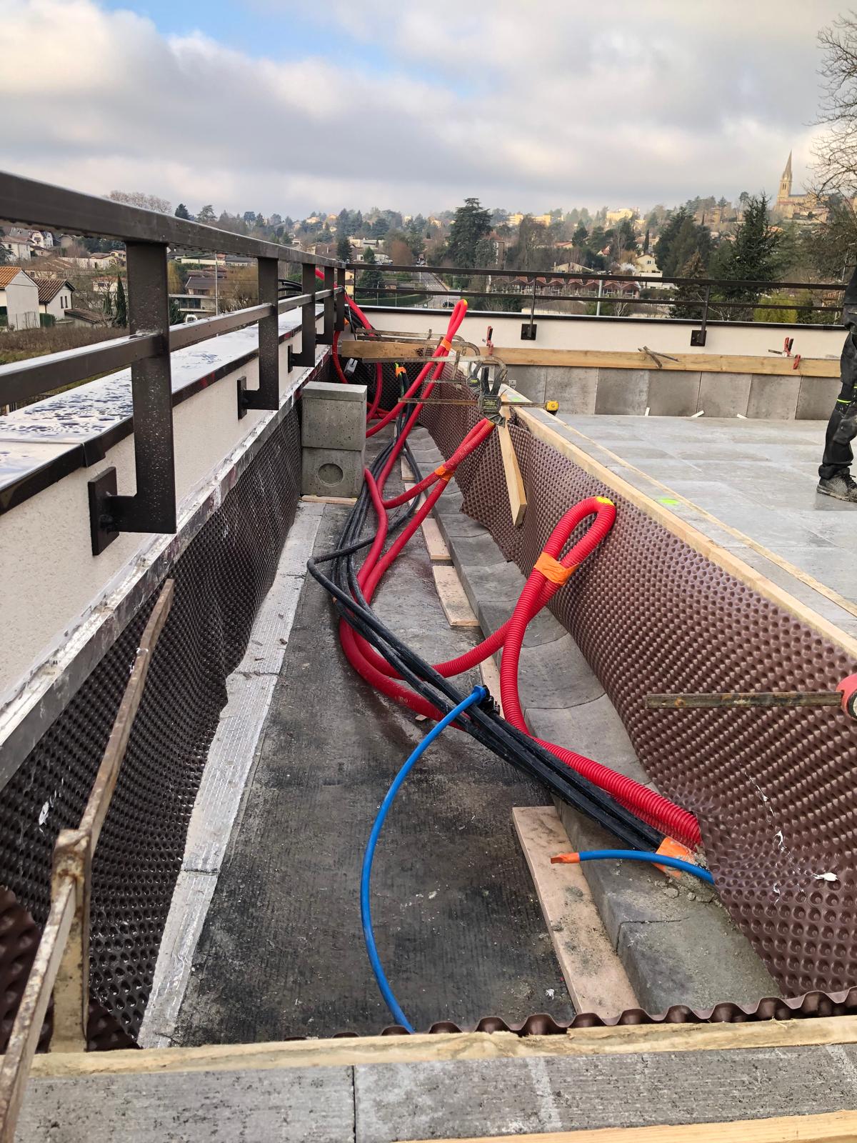 Chantier de terrasse en cours : gaines rouges et noires, drainage alvéolaire, et garde-corps en métal avec vue sur ville.