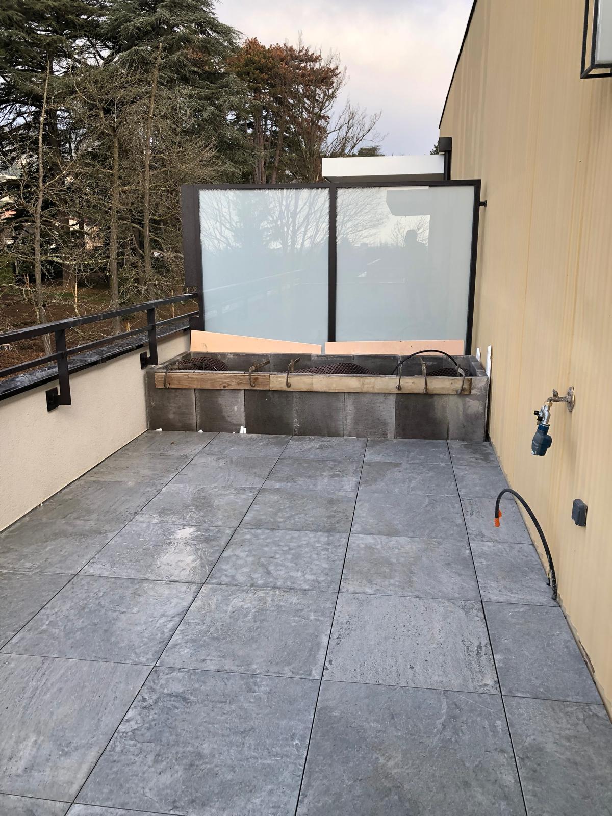 Terrasse extérieure en cours de finition avec dalles grises, garde-corps noir, structure de barbecue en parpaings et vue sur arbres nus.