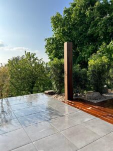 Terrasse moderne avec dalles grises humides et plancher bois brillant, entourée de verdure luxuriante sous un ciel bleu clair.