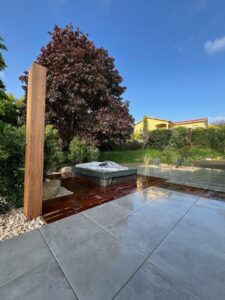 Espace détente extérieur moderne avec jacuzzi bâché, terrasse bois humide, carrelage gris clair et grand arbre rouge sous un ciel bleu vif.