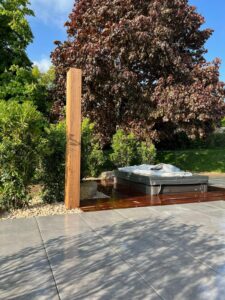 Terrasse moderne avec jacuzzi foncé sur plancher bois rougeoyant, entouré de verdure et d'un pilier bois, sous ciel bleu.