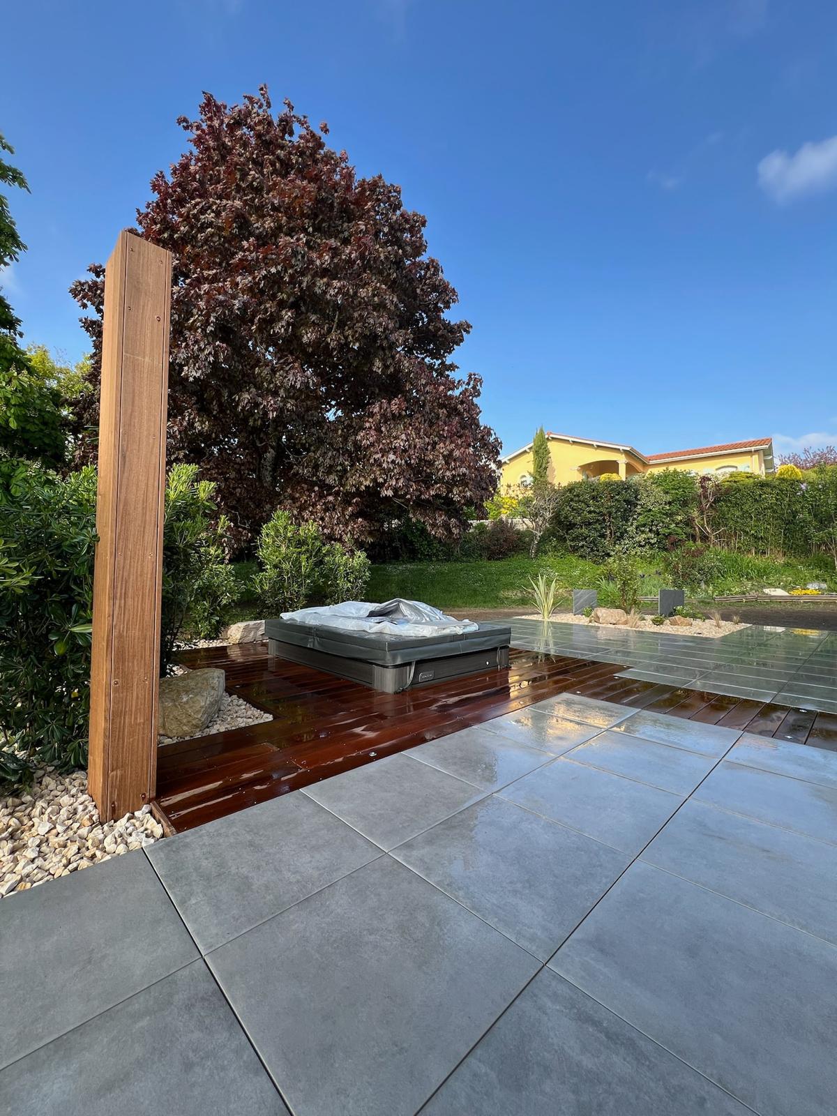 Espace détente extérieur moderne avec jacuzzi bâché, terrasse bois humide, carrelage gris clair et grand arbre rouge sous un ciel bleu vif.