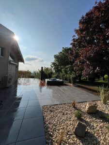 Terrasse moderne carrelée gris brillant et mouillée, avec jacuzzi sur platelage bois, sous un soleil éclatant et ciel bleu.