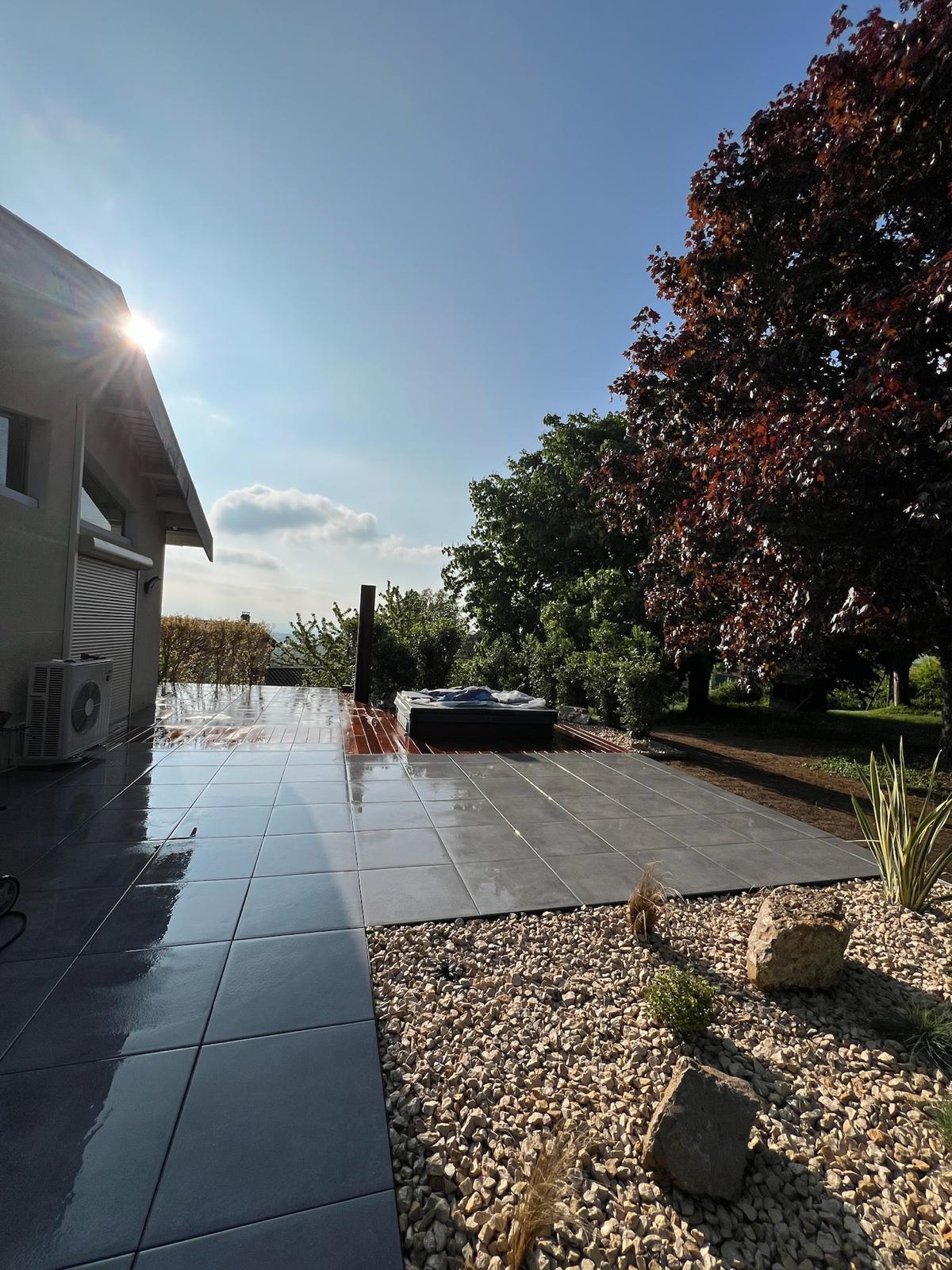 Terrasse moderne carrelée gris brillant et mouillée, avec jacuzzi sur platelage bois, sous un soleil éclatant et ciel bleu.