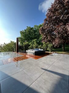 Terrasse moderne avec douche extérieure et jacuzzi, dalles grises et bois mouillé sous un ciel bleu clair.