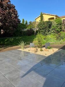 Terrasse moderne carrelée gris brillant à côté d'un parterre de gravier clair, plantes et rochers, devant une pelouse et maison jaune.