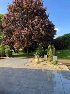 Grande terrasse en dalles grises brillantes juxtaposée à un parterre de gravier clair, dominé par un érable rouge et un ciel bleu clair.