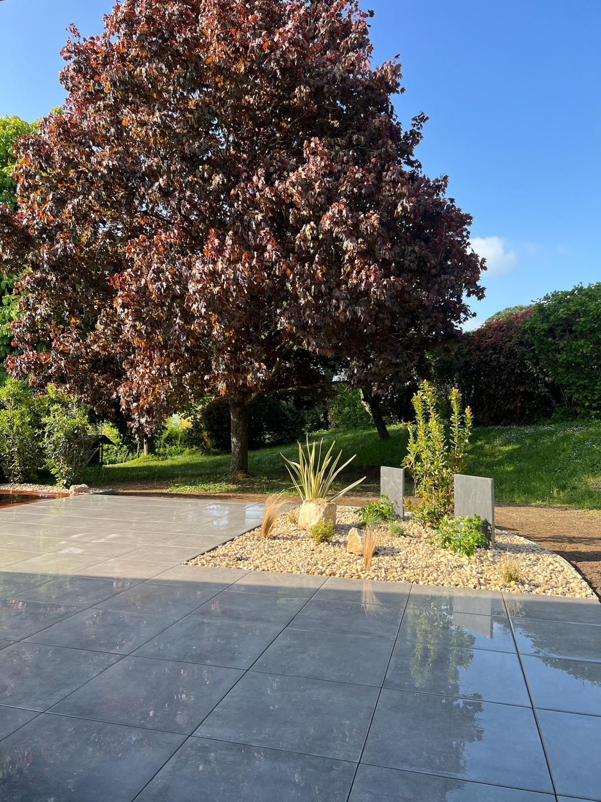 Grande terrasse en dalles grises brillantes juxtaposée à un parterre de gravier clair, dominé par un érable rouge et un ciel bleu clair.
