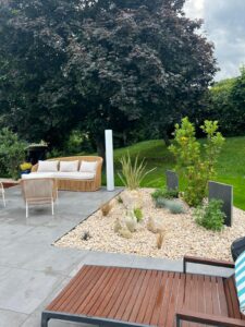 Salon extérieur moderne avec canapé en rotin, mobilier contemporain, et jardin sec en galets et plantes grasses sous un grand arbre foncé.