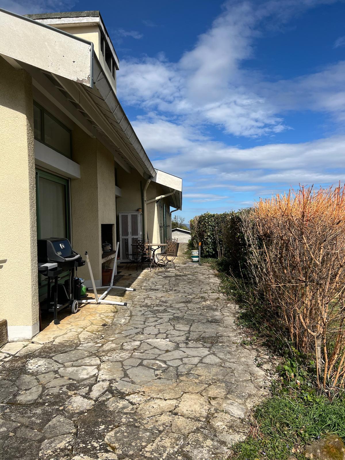 Terrasse dallée de pierres inégales longeant une maison jaune pâle, avec un barbecue et un salon de jardin sous un ciel bleu et nuageux.