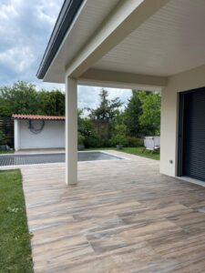 Terrasse carrelée effet bois sous auvent, donnant sur une piscine couverte et un jardin verdoyant par temps nuageux.