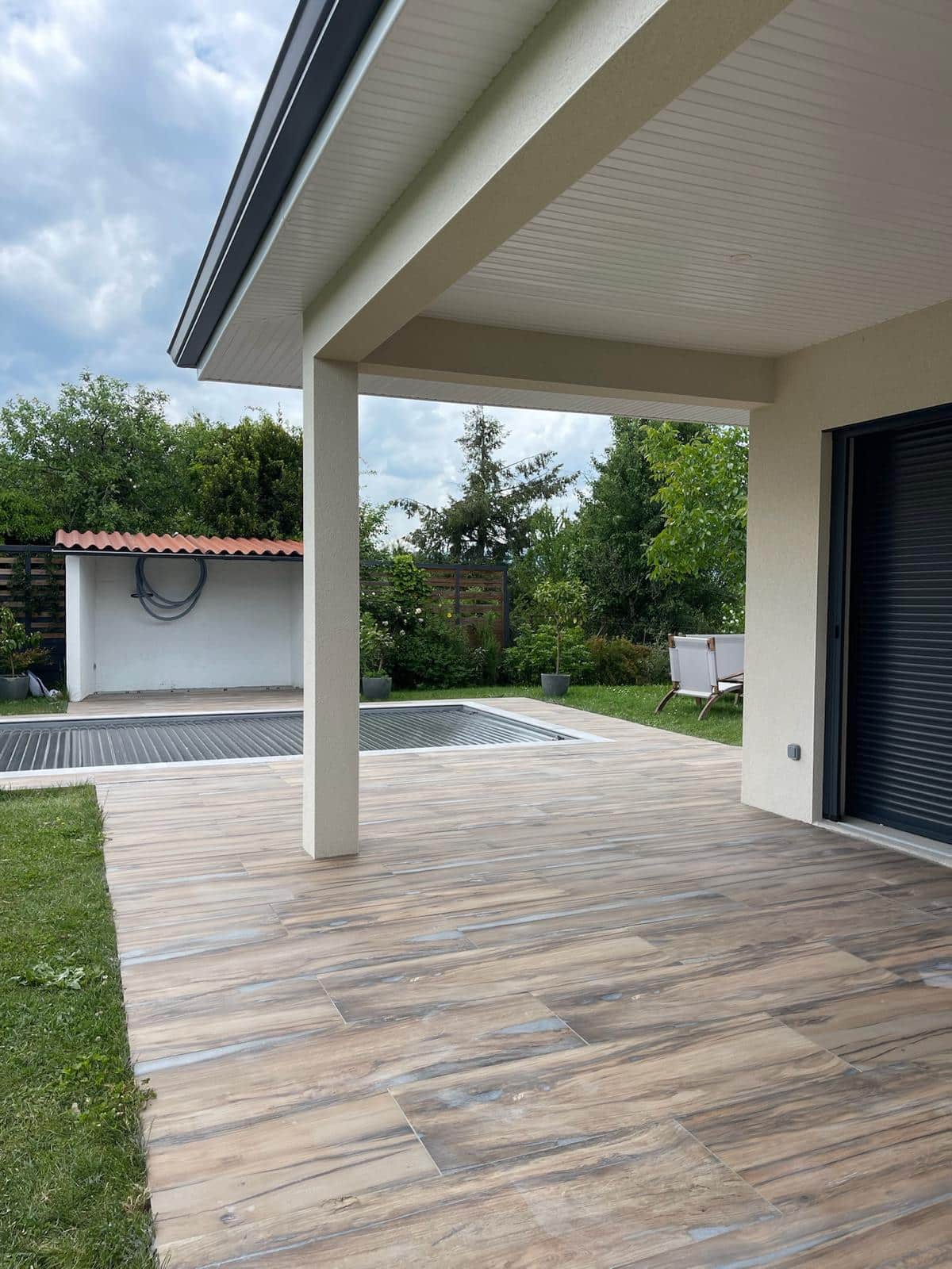 Terrasse carrelée effet bois sous auvent, donnant sur une piscine couverte et un jardin verdoyant par temps nuageux.