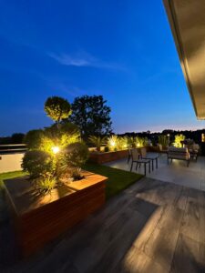 Terrasse moderne au crépuscule avec éclairage chaleureux mettant en valeur des jardinières en bois et des arbres taillés.