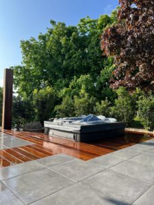 Spa extérieur noir couvert, posé sur une terrasse en bois brillant juxtaposée à des dalles de carrelage grises, entouré d'une végétation luxuriante.