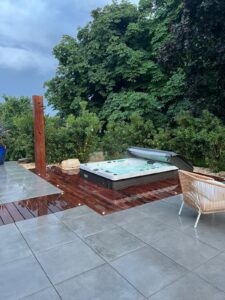 Spa extérieur sur terrasse bois rouge et carrelage gris, sous un ciel nuageux, entouré de verdure dense.