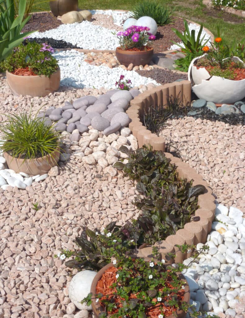 Jardin paysager moderne avec allée de galets gris, graviers blancs et roses, bordures, et plantes en pots fleuris violets et oranges.