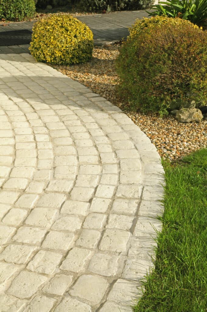 Chemin pavé clair et courbé dans un jardin, bordé d'herbe verte et de gravier avec des arbustes colorés, menant à une terrasse en bois.