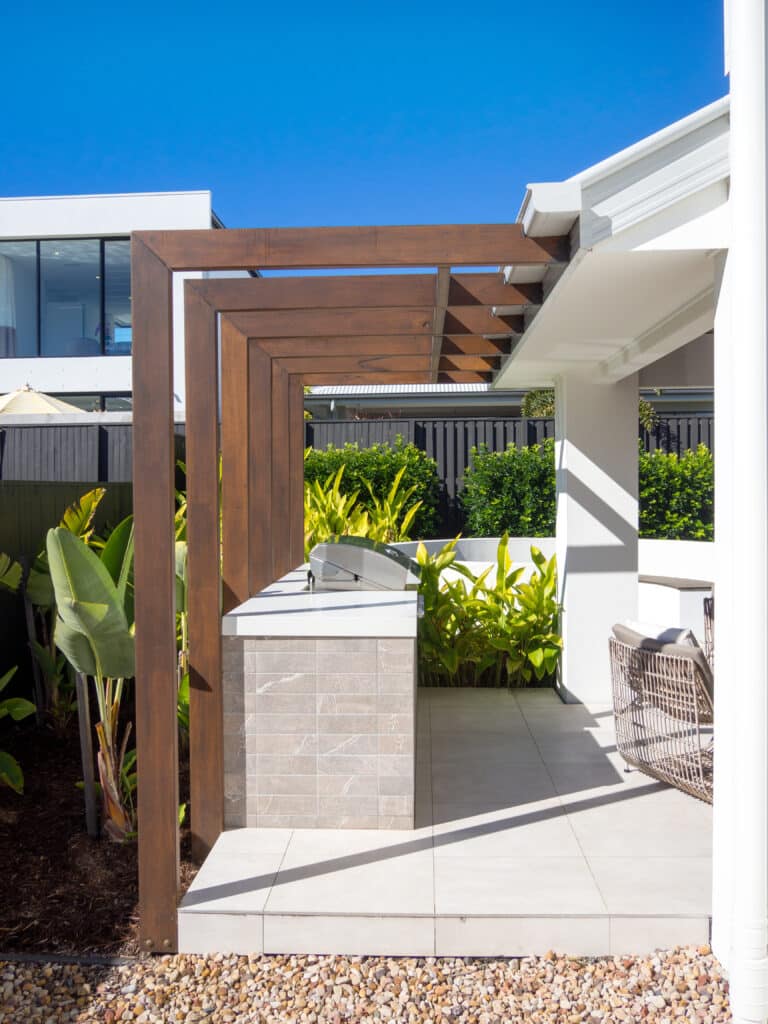 Pergola bois et cuisine extérieure moderne avec barbecue intégré, patio carrelé clair, végétation luxuriante et ciel bleu.