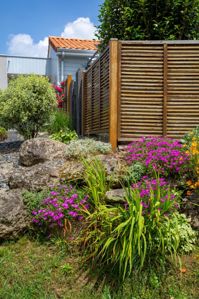 Jardin rocailleux coloré avec fleurs violettes, feuillage vert, grosses pierres, clôture en bois, maison à toit de tuiles sous ciel bleu.