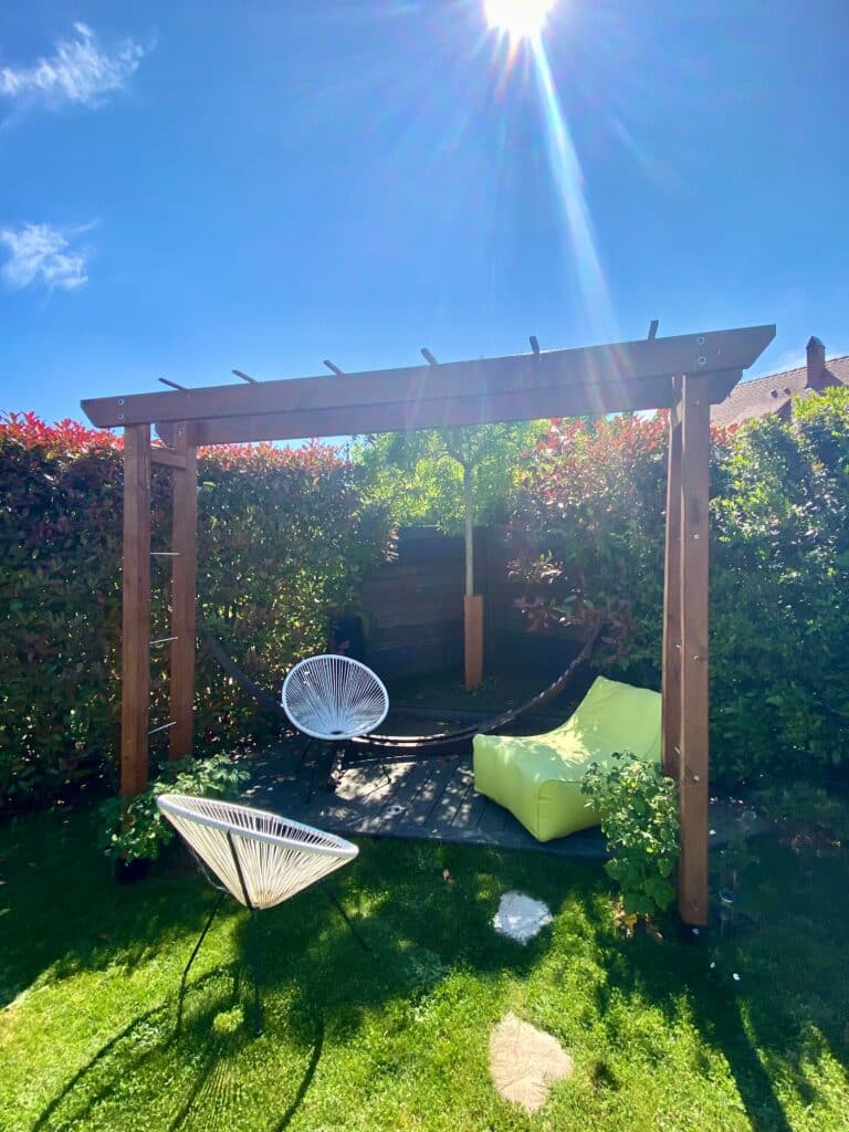 Pergola en bois dans un jardin ensoleillé avec deux chaises Acapulco blanches et un pouf vert sur terrasse. Ambiance extérieure lumineuse et relaxante.