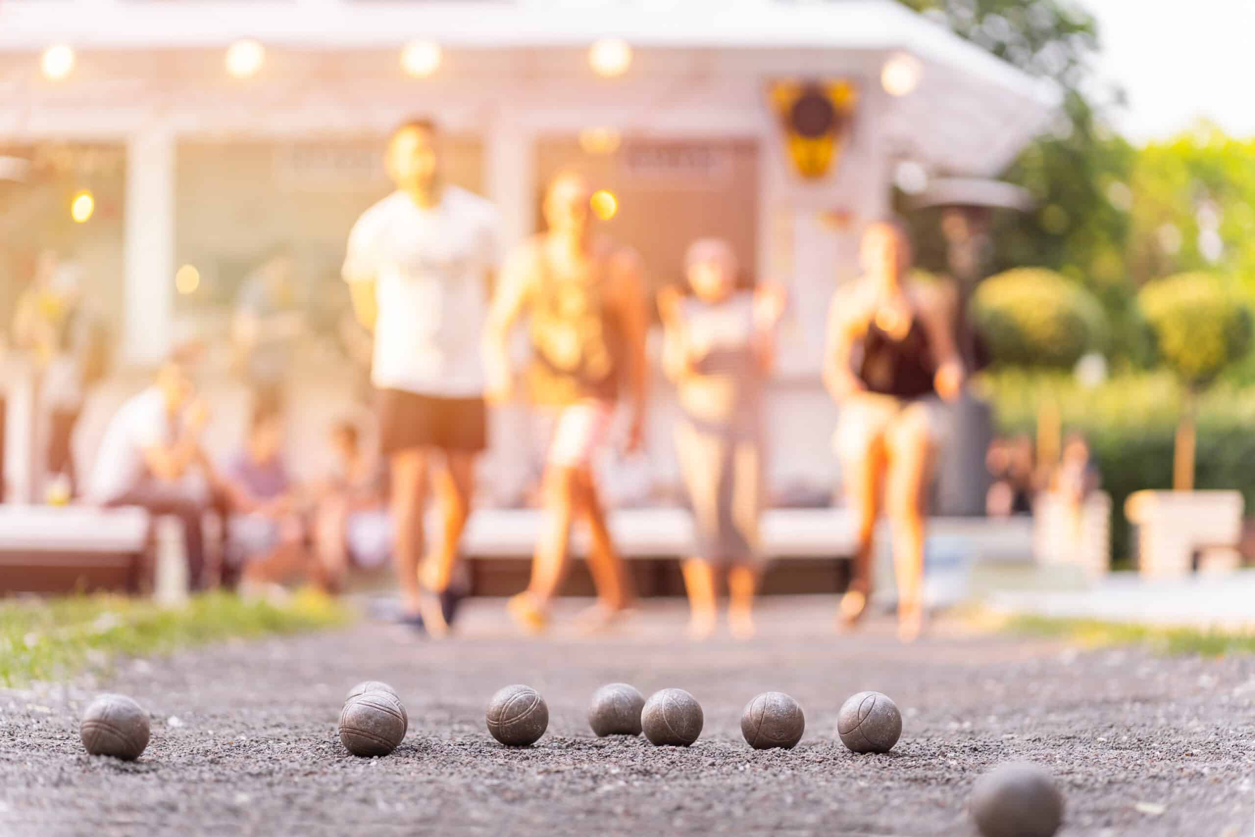Boules de pétanque grises sur un terrain de gravier, personnes floues et lumières chaudes en arrière-plan, jouant ou se détendant en extérieur.
