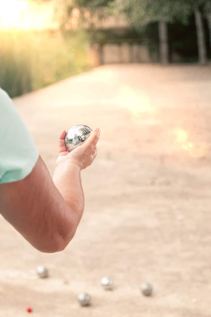 Bras d'un joueur tenant une boule de pétanque brillante, prêt à lancer sur un terrain ensoleillé. D'autres boules et le cochonnet sont au sol.