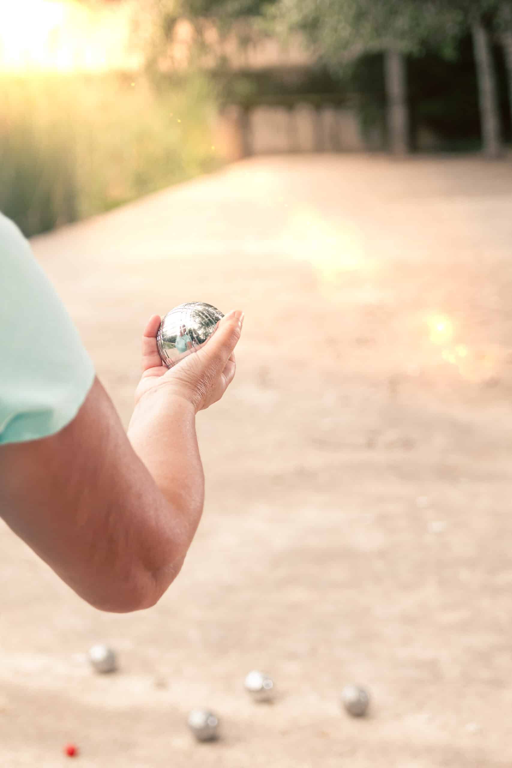 Bras d'un joueur tenant une boule de pétanque brillante, prêt à lancer sur un terrain ensoleillé. D'autres boules et le cochonnet sont au sol.