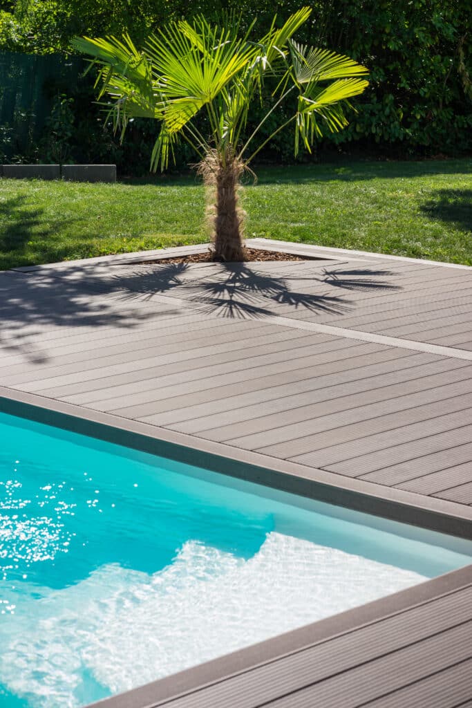 Piscine bleue, margelle en bois composite et palmier. Ses feuilles vertes projettent des ombres sur la terrasse et le jardin ensoleillé.