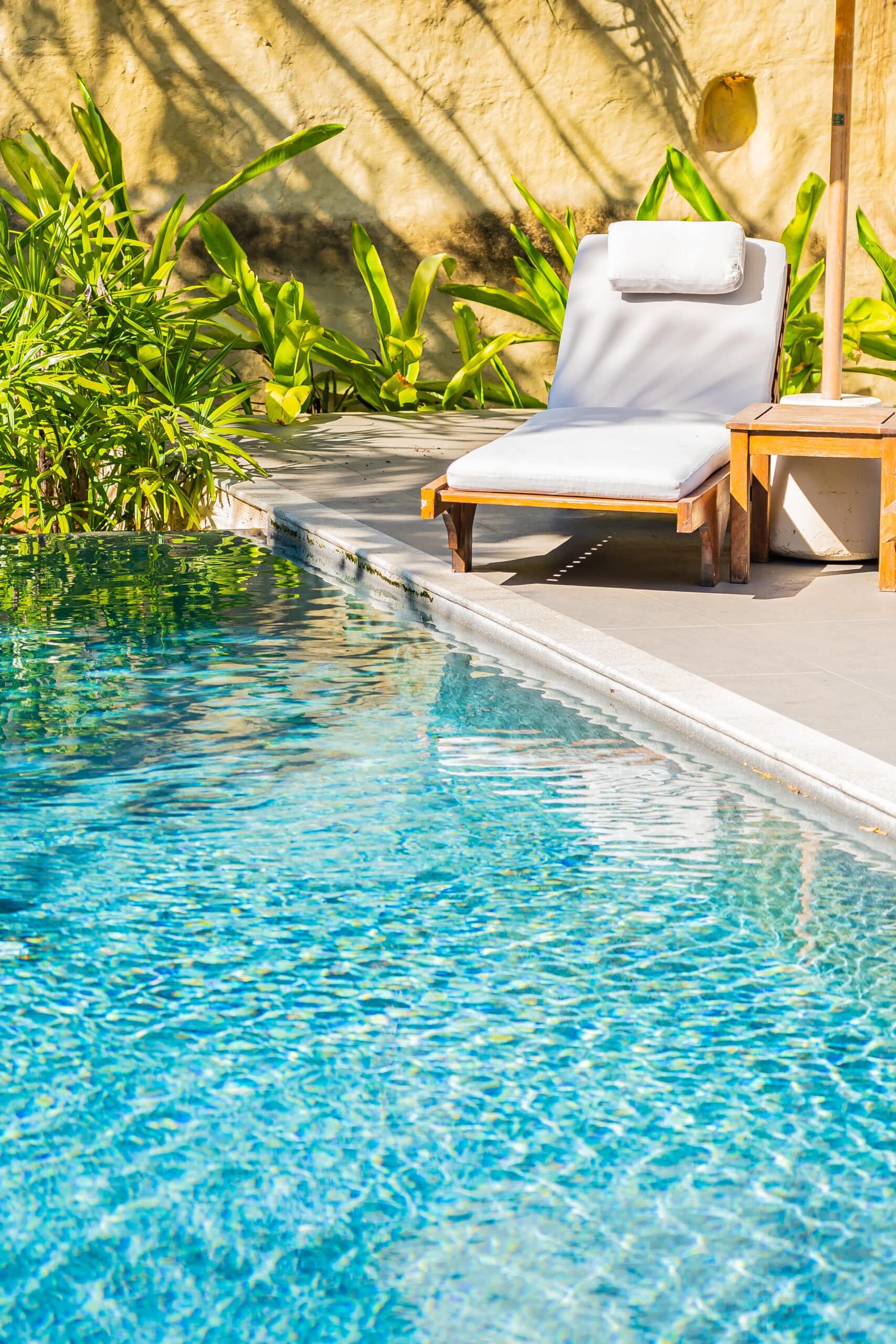 Piscine bleue scintillante avec transat en bois et coussin blanc, bordée de plantes tropicales luxuriantes et d'un mur ocre au soleil.